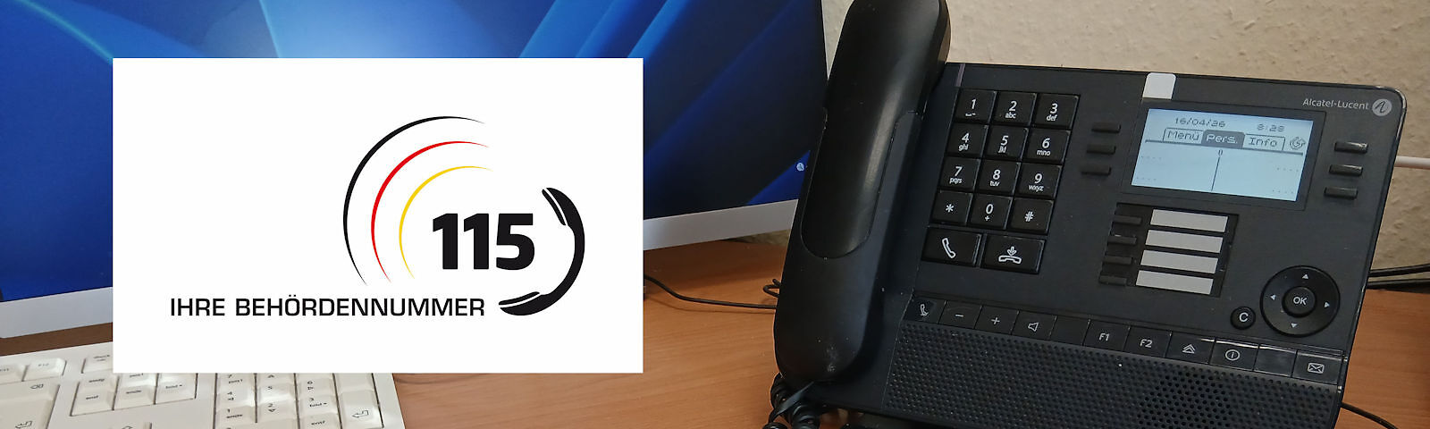 Schreibtisch mit Tastatur, Telefon und Bildschirm mit dem Logo der Behördennummer 115